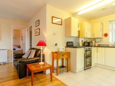 A kitchen area with wooden cabinets and appliances next to a living room with a black leather couch and a wooden side table at Brook Cottage in Rosecraddoc Lodge near Liskeard