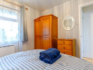A bedroom with a wooden wardrobe and chest of drawers beside a bed with striped bedding and folded towels at Brook Cottage in Rosecraddoc Lodge near Liskeard