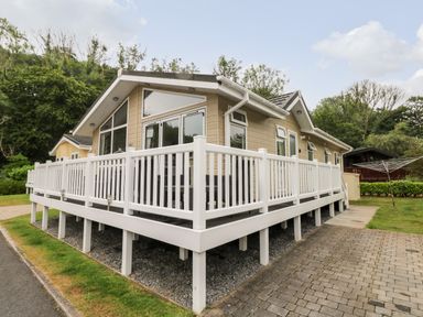 A beige cabin with large windows and a white railing around a raised deck next to a paved driveway at 95 The Haven in Wisemans Bridge near Saundersfoot
