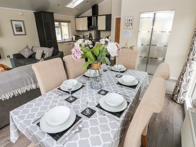 A dining table with six beige chairs set with white plates and glasses and a flower vase in a room with a kitchen and sofa at 95 The Haven in Wisemans Bridge near Saundersfoot