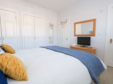 A bedroom with a bed covered with white and blue bedding yellow pillows white wardrobes a dresser with a television and a mirror on the wall at Captains Retreat in Christchurch