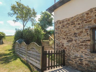A stone exterior wall of a building with a wooden gate and fence leading to steps surrounded by trees and grass at Owls Nest in South Molton