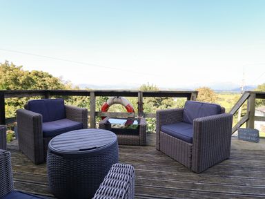 An outdoor deck with wicker chairs cushions a round table and a view of trees and hills at Snowdon View in Harlech