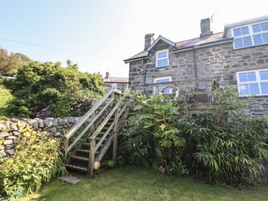 An exterior view of a stone house with a wooden staircase and garden plants at Snowdon View in Harlech