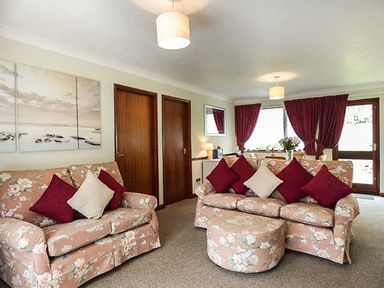 A living room with floral sofas and matching ottoman with red and cream cushions at Chy Lowenna Rosecraddoc Holiday Village near Liskeard