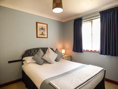 A bedroom with a double bed three pillows a framed picture on the wall bedside table lamp and window with curtains at Chy Lowenna Rosecraddoc Holiday Village near Liskeard