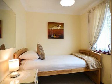 A small bedroom with a single wooden bed a bedside table with a lamp a painting on the wall and a window with curtains at Chy Lowenna in Rosecraddoc Holiday Village near Liskeard