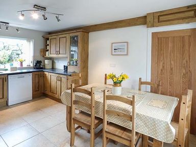 A kitchen with wooden cabinets dining table with tablecloth and chairs and flowers in a vase at Simdda Wen Cottage in Llanddona near Beaumaris