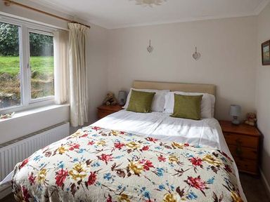 A bedroom with a double bed covered with a floral blanket two green pillows bedside tables with lamps and teddy bears and a window with curtains at Simdda Wen Cottage in Llanddona near Beaumaris