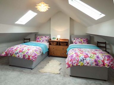 A bedroom with two single beds with floral bedspreads and blue throws a wooden nightstand with a lamp and baskets under skylights at Simdda Wen Cottage in Llanddona near Beaumaris