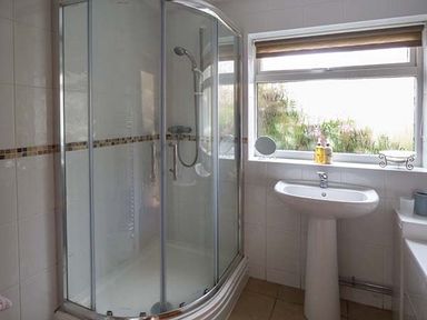 A bathroom with a curved glass shower enclosure a pedestal sink and a window with toiletries on the sill at Simdda Wen Cottage in Llanddona near Beaumaris