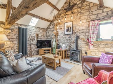 A living room with stone walls wooden beams a wood-burning stove a leather sofa and armchairs a television and wooden furniture at Berts Cottage in Heights near Appleby-in-Westmorland