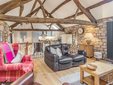 A living room with wooden beams stone walls leather sofa red plaid armchair and kitchen area at Berts Cottage Heights near Appleby-in-Westmorland