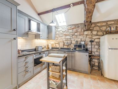 A kitchen with gray cabinets, black countertop, stone wall, kitchen island, stove, oven, microwave, and refrigerator at Berts Cottage in Heights near Appleby-in-Westmorland