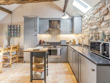 A kitchen with grey cabinets a stone wall a wooden table with chairs and a portable kitchen island at Berts Cottage in Heights near Appleby-in-Westmorland
