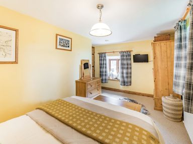 A bedroom with a bed a wooden dresser a window with plaid curtains and a wall-mounted television at Berts Cottage Heights near Appleby-in-Westmorland
