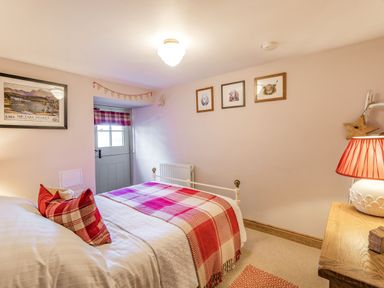 A bedroom with a bed covered by a red and white checked blanket and pillows a wooden desk with a lamp a door and framed pictures on the walls at Berts Cottage in Heights near Appleby-in-Westmorland