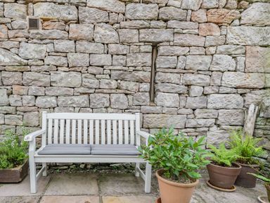 A white bench with gray cushions in front of a stone wall with potted plants on a paved floor at Berts Cottage Heights near Appleby-in-Westmorland