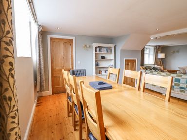 A dining room with a wooden table surrounded by chairs next to a living area with sofas and a bookshelf at Is Y Deri in Harlech