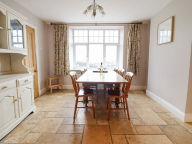 A dining room with a wooden table and six chairs near a window with floral curtains at Is Y Deri in Harlech