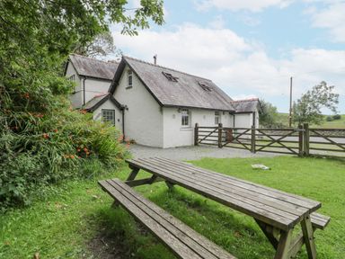 A wooden picnic table on grass next to a white house with a tiled roof and wooden gates at Is Y Deri in Harlech