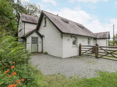 An exterior view of a white house with a slate roof gravel driveway wooden gate and surrounding greenery at Is Y Deri in Harlech