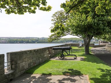 A riverside walkway with a historic cannon on grass near a stone wall and trees at Iona 10 Palace Street East in Berwick-upon-Tweed