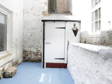 A small outdoor area with white stone walls a white door blue floor and a hanging flower basket at Iona 10 Palace Street East Berwick-upon-Tweed