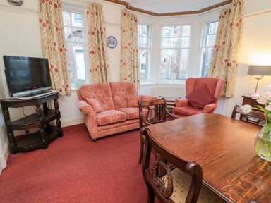 A seating area with a pink sofa and armchair next to a bay window with floral curtains a wooden dining table with chairs and a television on a stand in Iona 10 Palace Street East Berwick-upon-Tweed
