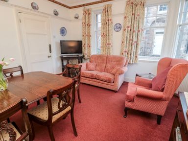 A living room with a wooden dining table and chairs a television on a stand a two-seater sofa an armchair and windows with patterned curtains at Iona 10 Palace Street East in Berwick-upon-Tweed