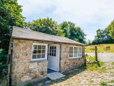 A stone cottage with white window frames and a half-open door surrounded by trees and grass at Treverbyn Smithy in St Neot