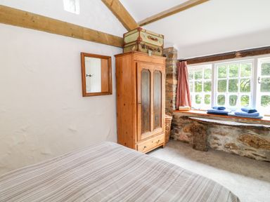 A bedroom with a wooden wardrobe, mirror on the wall, window with folded towels on the sill and stone wall at Treverbyn Smithy in St Neot