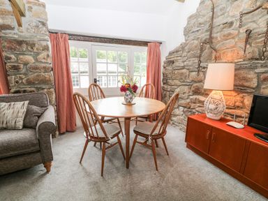 A dining area with a round wooden table and four wooden chairs near a window with red curtains at Treverbyn Smithy in St Neot