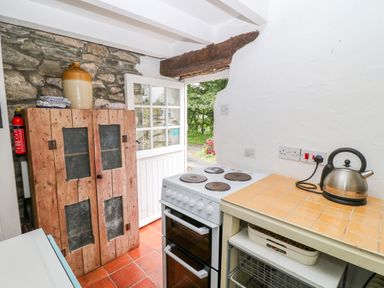 A kitchen with a stove a kettle on a tiled counter and a wooden cabinet against a stone wall at Treverbyn Smithy in St Neot