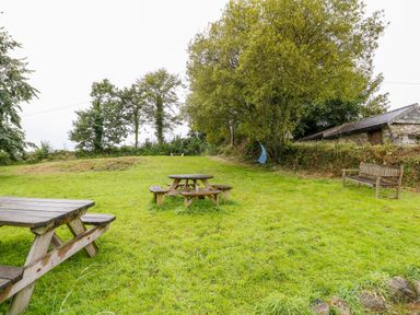 A grassy outdoor area with wooden picnic benches a wooden bench a tree with a hanging blue swing and a stone building at Treverbyn Smithy in St Neot