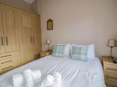 A bedroom with a bed with white linens and plaid cushions flanked by wooden bedside tables with lamps and a wooden wardrobe at 22 Thirlmere in Troutbeck Bridge