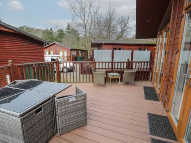 An outdoor patio with rattan chairs a small table and a folded rattan table set near wooden cabins at 22 Thirlmere in Troutbeck Bridge