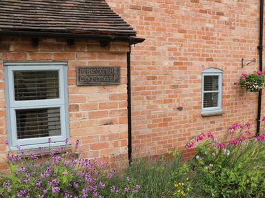 A brick wall of Pebworth Cottage with two windows flowers and a hanging flower basket at Pebworth near Stratford-upon-Avon