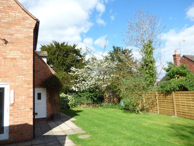 A garden with a lawn trees and shrubs next to a brick house with a tiled patio at Pebworth Cottage in Pebworth near Stratford upon Avon