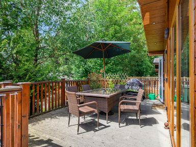 An outdoor wooden deck with a glass dining table, six wicker chairs, a large umbrella, a barbecue grill, and surrounding trees at Claife View Lodge in Troutbeck Bridge