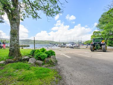 A lakeside view with boats docked at a marina a tractor on a paved road and a large tree with grass and plants nearby at Claife View Lodge in Troutbeck Bridge