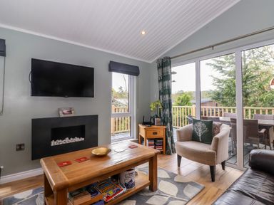 A living room with a wall mounted TV fireplace wooden table armchair and sliding glass doors at Claife View Lodge in Troutbeck Bridge