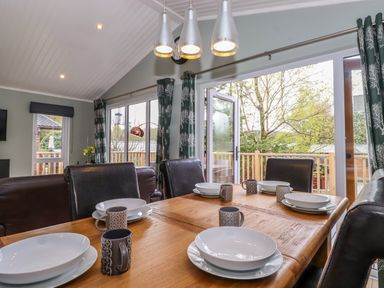 A dining area with a wooden table set with white bowls and patterned mugs and leather chairs near glass doors with curtains at Claife View Lodge in Troutbeck Bridge