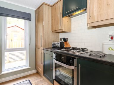 A kitchen with wooden cabinets a gas stove an oven a knife block a toaster and a window showing a wooden shed outside at Claife View Lodge in Troutbeck Bridge