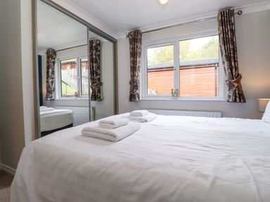 A bedroom with a bed covered in white linens towels on the bed window with patterned curtains and a mirrored wardrobe at Claife View Lodge in Troutbeck Bridge
