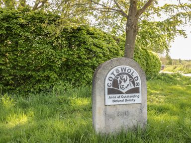 A stone marker with a sheep illustration and text about Cotswolds area of outstanding natural beauty next to a tree and hedge