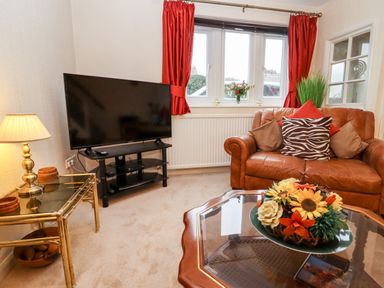A living room with a leather sofa cushions a television on a stand and a glass coffee table with a flower arrangement at Rosemary Cottage in Bourton-on-the-Water