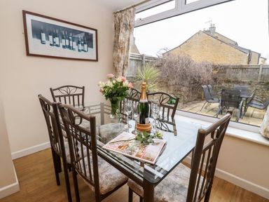 A dining room with a glass table and six chairs with a bouquet of flowers and a champagne bottle beside glasses and magazines at Rosemary Cottage in Bourton-on-the-Water