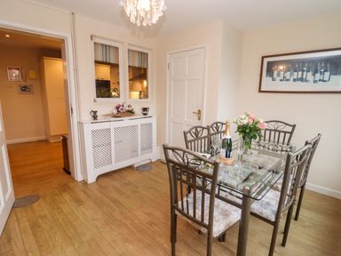 A dining room with a glass table and six chairs a flower vase and a champagne bottle on the table a white cabinet and a framed picture on the wall at Rosemary Cottage in Bourton-on-the-Water