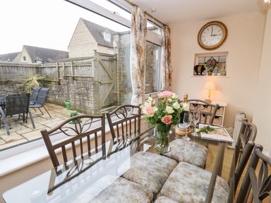 A dining area with a glass table flowers on the table and chairs next to a window overlooking a patio at Rosemary Cottage in Bourton-on-the-Water